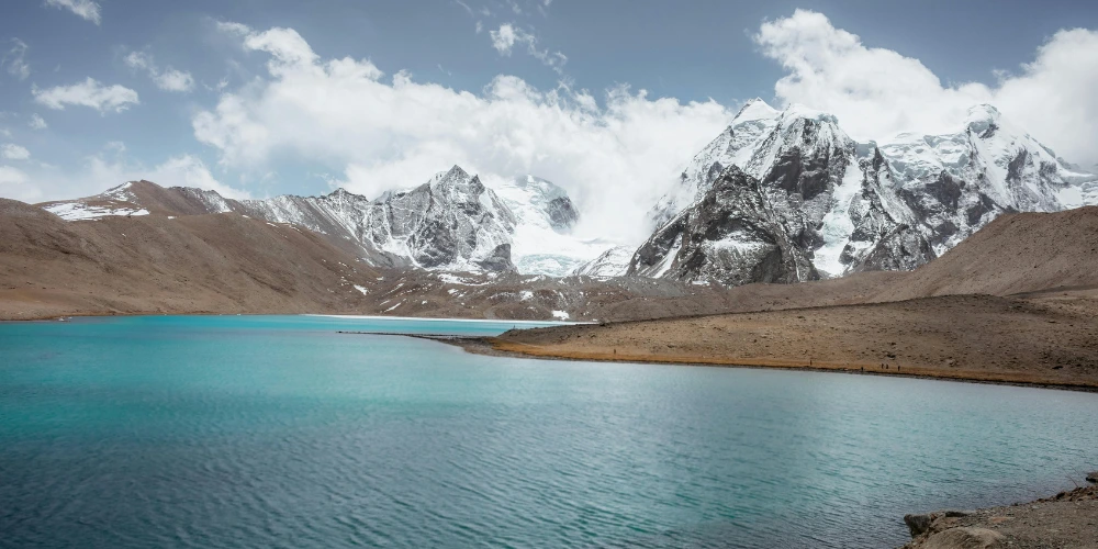 A spectacular mountain panorama of Sikkim featuring a tourist vehicle on a winding road with travelers, vibrant Buddhist prayer flags, a traditional monastery nestled in the hills, and a turquoise glacial lake under the snow-capped Himalayan peaks.